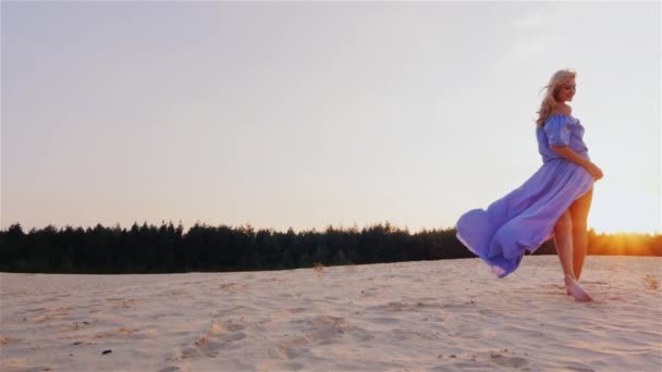 Una mujer delgada con un vestido azul claro de pie en la playa al atardecer. El viento juega con su vestido — Vídeos de Stock