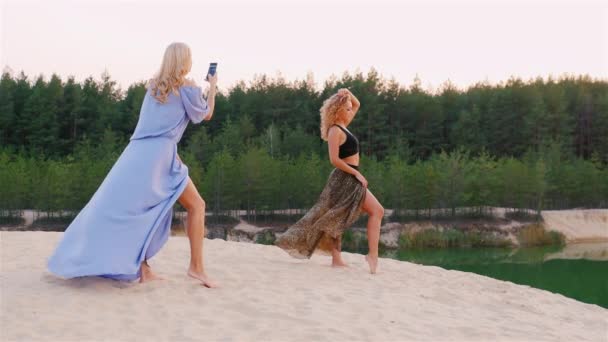 Belle femme en robe posant teint sa petite amie, qui l'a photographiée au téléphone. Sur le lac pittoresque dans une forêt — Video