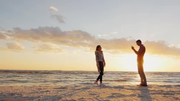 若いカップルは日没時に海で撮影されます。ガールフレンドの電話を撮影した男ボーイフレンドのためにポーズをとる女性 — ストック動画