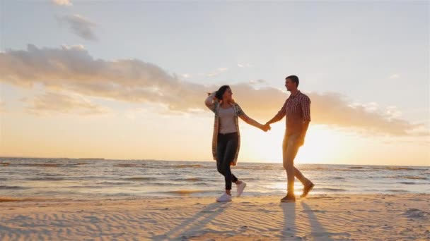 Steadicam slow motion shot: Romantic couple walking on the beach at sunset, holding hands, having a good time — Stock Video