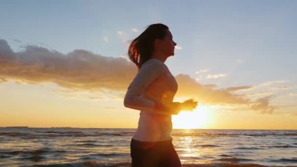 Mujer joven con una hermosa figura corriendo en la playa al atardecer. El pelo largo es hermoso revoloteando en el viento. Steadicam toma de cámara lenta — Vídeos de Stock