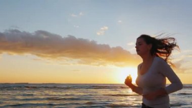 Çekici genç kadın gün batımında deniz kıyısında koşuyor. Spor - sağlıklı yaşam tarzı meşgul. Steadicam yavaş çekim atış