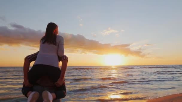 Young couple in love having fun on the beach at sunset berugi. A girl sits on the shoulders of men running on the beach, laughing. Honeymoon, the energy of youth, love. Steadicam slow motion shot — Wideo stockowe