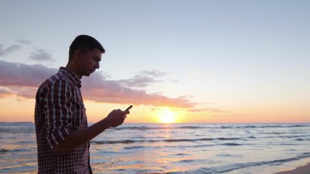 Jovem solitário caminhando ao longo da praia ao pôr do sol, digitando uma mensagem no telefone — Vídeo de Stock