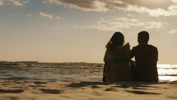 Una pareja amorosa sentada en la playa al atardecer. Día fresco, vista trasera — Vídeos de Stock