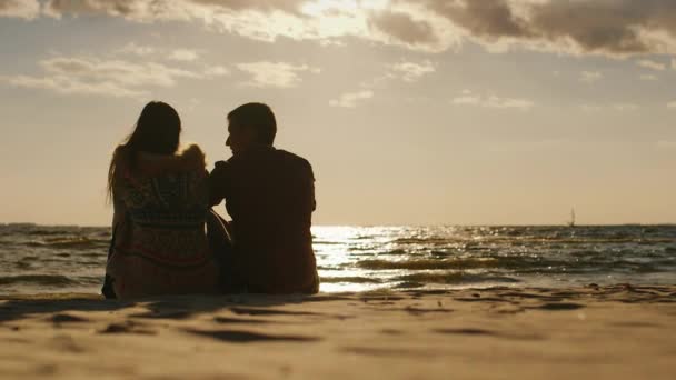 Siluetas de parejas enamoradas, sentadas en la playa, mirando el atardecer y una tabla con vela. Vista trasera — Vídeo de stock