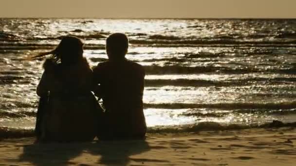 Siluetas de parejas enamoradas, sentadas sobre un fondo del mar, que refleja el sol poniente, vista trasera — Vídeo de stock