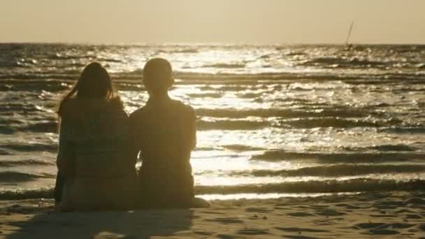 Pareja joven enamorada sentada en la playa, observando el atardecer y la persona que monta en windsurf. Vista trasera — Vídeo de stock