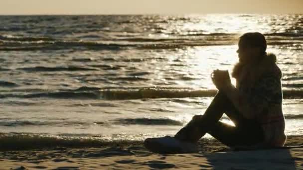 Jovem mulher sentada na praia em um dia ventoso fresco, bebendo uma bebida quente de um copo. Mantenha-se quente no tempo frio. Ao pôr do sol — Vídeo de Stock