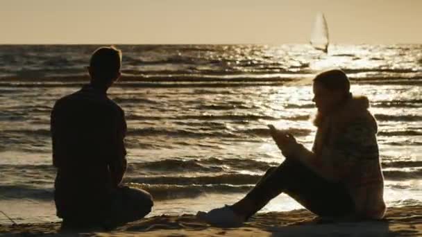 Casal romântico relaxante na praia ao pôr do sol. Mulher desfrutando de um telefone, um homem joga pedras na água. Nas velas distantes com uma placa de vela — Vídeo de Stock