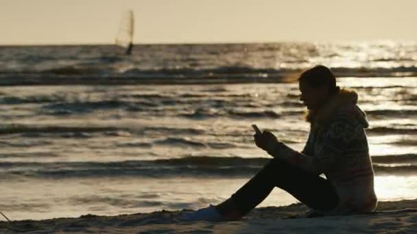 Силуэт молодой женщины на берегу моря, сидящей на песке, наслаждающейся телефоном. На заднем плане закат и доска для плавания с саилом — стоковое видео