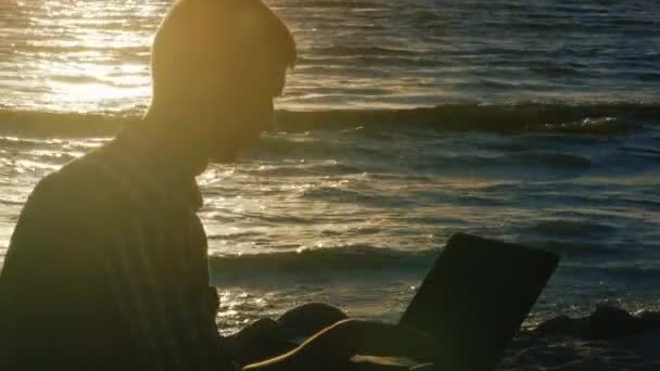 Joven trabajando con un portátil junto al mar al atardecer — Vídeo de stock