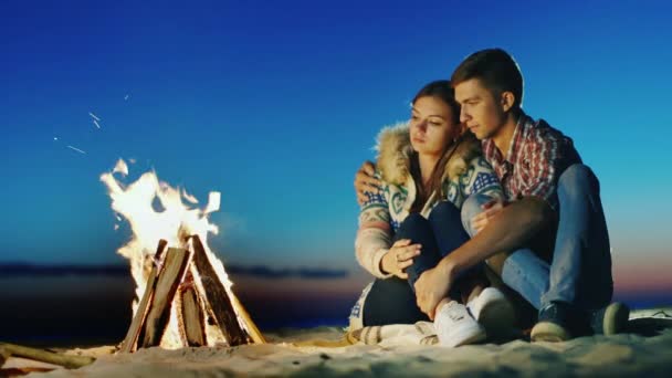 Romántica pareja joven sentada en la fogata de la playa. Abrazo, admirando el fuego, soñando — Vídeos de Stock