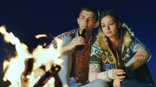 Fiatal pár lazít a tűz mellett, söröznek vagy isznak az üvegből. — Stock videók