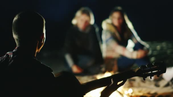 A companhia de amigos relaxando junto ao fogo, um homem toca guitarra — Vídeo de Stock