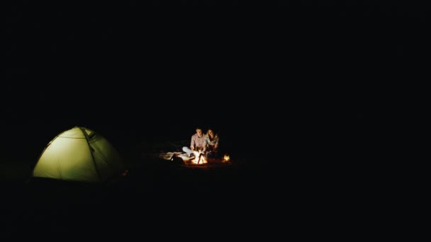 Вид сверху на молодую пару, палатку и костер. Вокруг темно, только огонь и палатка освежить людей — стоковое видео