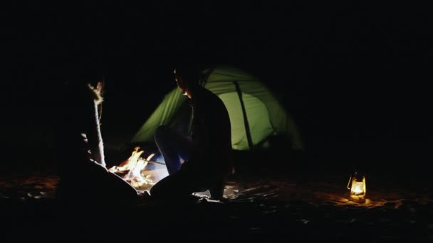 对年轻的夫妇烤棉花糖棒周围的篝火和在夜里照亮的帐篷。后视图 — 图库视频影像