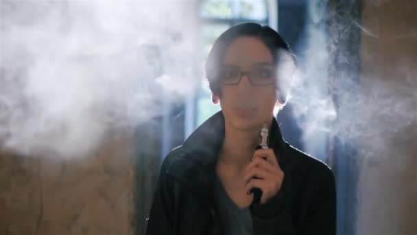 Une jeune gothique fume une cigarette électronique contre la fenêtre. Fumée arrière magnifiquement éclairée par la lumière — Video