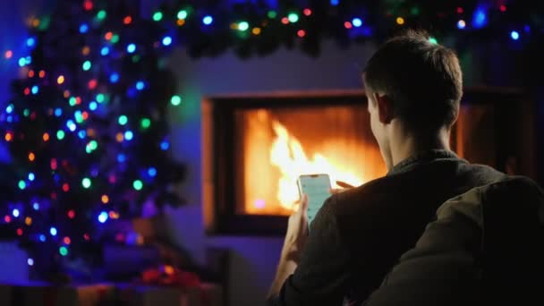 Joven utiliza un teléfono inteligente junto a la chimenea decorada para Navidad. Orden de regalo — Vídeos de Stock