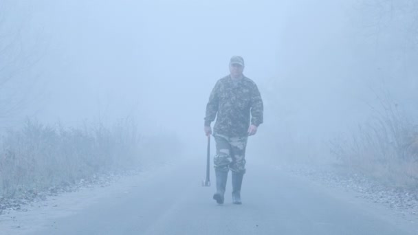 Extraño hombre con un hacha camina por el camino en la niebla. — Vídeos de Stock