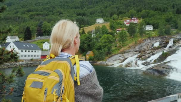 Uma mulher admira a aldeia norueguesa e a cachoeira. Viajar na Escandinávia — Vídeo de Stock