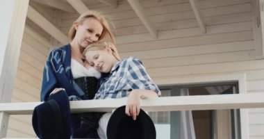 Anne ve kızı ellerinde şapkalarla ahşap bir evin verandasında duruyorlar.