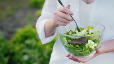Beyaz gömlekli bir kadın taze salata yiyor, büyüdüğü yatağın yanında duruyor.