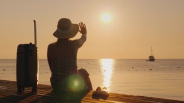 Bir kadın uzaktaki bir gemiye elini sallıyor, yol çantasındaki iskelede oturuyor.