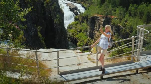 Frau fotografiert sich vor dem Hintergrund des Wasserfalls von Woringsfossen in Norwegen. Beeindruckende Schönheit der skandinavischen Natur — Stockvideo
