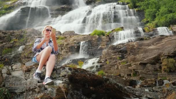 Um viajante usa um smartphone contra o fundo de uma cachoeira Tvindefossen na Noruega — Vídeo de Stock