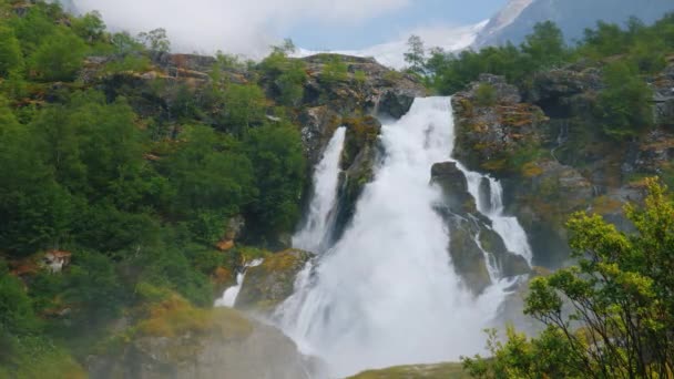 Hermosa cascada de las aguas del glaciar. Al fondo, la montaña con hielo en la parte superior es el glaciar Briksdal. La naturaleza de Noruega y Escandinavia — Vídeos de Stock