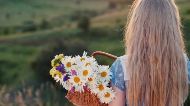 Bakifrån: Ett barn håller en korg med blommor, står på en pittoresk plats mot bakgrund av ängar och gröna kullar — Stockvideo