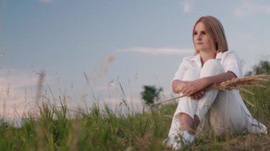 Beyaz elbiseli bir kadın sıcak bir yaz akşamında yeşil bir tepede dinleniyor.