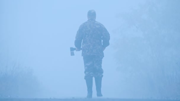 Un hombre con un hacha camina por el camino en la niebla - una escena terrible — Vídeo de stock