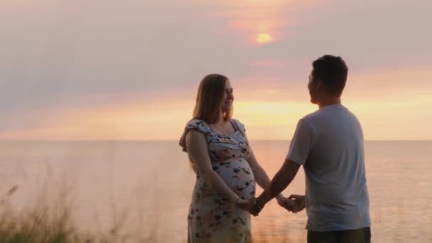 海に沈む夕日を背景に、多民族のモルトカップルが手をつないでいます。アジア人男性と彼の妊婦 — ストック動画