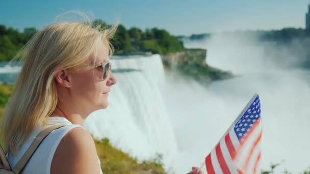 Porträt einer Frau vor dem Hintergrund der Niagarafälle in den USA, hält die amerikanische Flagge in der Hand — Stockvideo