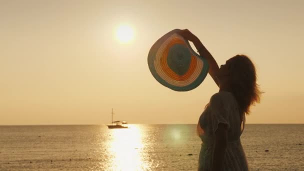 Uma mulher feliz encontra o amanhecer junto ao mar. Emocionalmente acenando seu chapéu, o navio é visto à distância — Vídeo de Stock