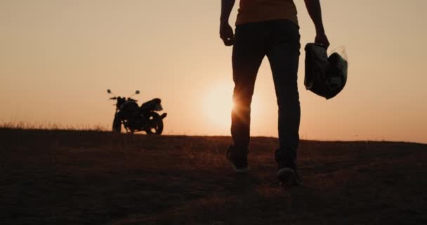 Die Silhouette eines Motorradfahrers mit einem Helm in der Hand, geht bei Sonnenuntergang zu seinem Motorrad — Stockvideo