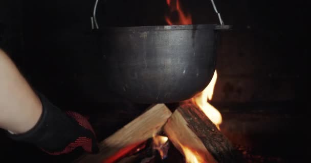 Una olla cuelga en la estufa de leña, el cocinero pone el tronco en el fuego — Vídeos de Stock