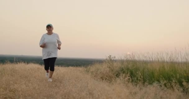 Uma mulher idosa corre ao longo de uma estrada rural, entra para o desporto. Visão traseira. Idosos activos — Vídeo de Stock