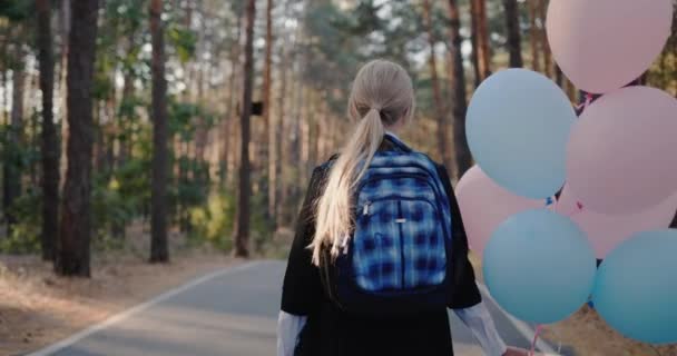 Skolflicka med en flock och ballonger promenader på vägen i parken med höga tallar — Stockvideo