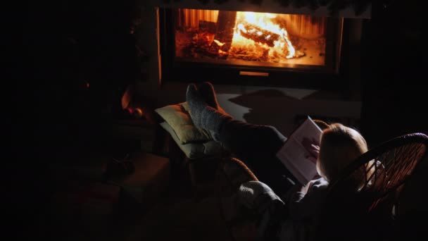 Mujer joven lee un libro mientras está sentada junto a la chimenea — Vídeos de Stock