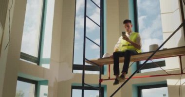 Yatalak bir işçi iskeleye kasksız oturur, akıllı telefonuyla oynar. Tehlikeli davranış konsepti