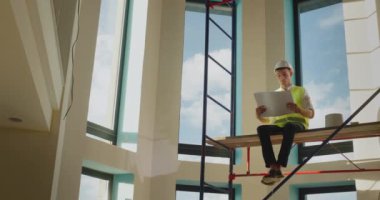 Genç bir işçi çizim üzerine çalışır, iskeleye dik oturur.