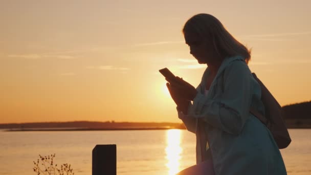 Una donna in un cappuccio si siede su una recinzione vicino a un pittoresco lago, utilizza uno smartphone. Bellissimo tramonto — Video Stock