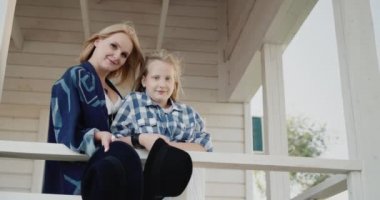 Anne ve kızı ellerinde şapkalarla ahşap bir evin verandasında duruyorlar.