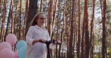 Parkta elektrikli scooter kullanan bir kız, mobiletin arkası balonlarla görülebilir.