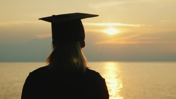 Uma mulher em um manto de graduação e em um boné olha para o nascer do sol sobre o mar — Vídeo de Stock