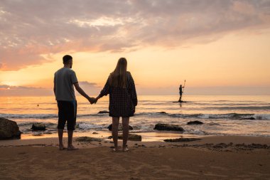Bulgaristan 'ın Varna kentindeki Poseidon heykelinin yanındaki kumsalda el ele tutuşan çift, sakin deniz ve altın gökyüzünü birlikte izliyor. Yüksek kalite fotoğraf