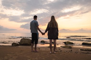 Çift el ele tutuşup sakin denizin üzerinde güneşin doğuşunu seyrediyor, kumlu bir sahilde çıplak ayakla duruyor ve sabah ışığı suya yansıyor. Yüksek kalite fotoğraf